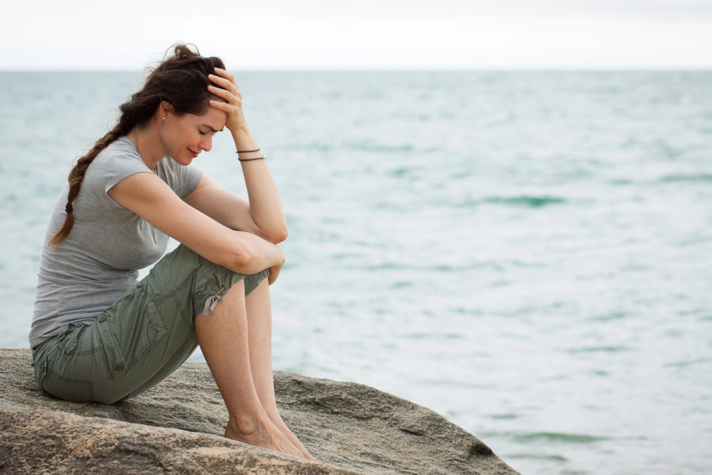 Woman sitting on a rock near ocean with hand on her forehead. She appears sad, as in grief.